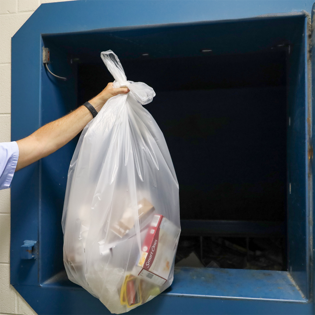 a UK student holding a clear bag of recyclables at an open compactor door.